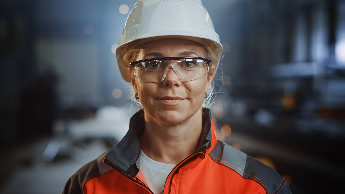 Eine Frau im Alter von 30 bis 40 Jahren. Sie trägt eine Schutzbrille und einen weißen Sicherheitshelm. Als Kleidung trägt sie eine orangene Sicherheitsjacke. Im Hintergrund sieht man das Gelände einer Chemiefabrik.